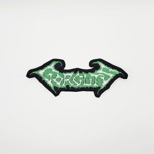 Puede incluir: Parche bordado con la palabra "FORGOTTEN" en verde, con letras estilizadas. El parche tiene un borde negro y una forma simétrica de flecha. El parche está sobre un fondo blanco.