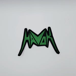 May include: An embroidered patch featuring the word "HAVAH" in green, stylized lettering with a black outline. The patch is set against a white background.