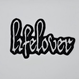 Könnte beinhalten: Schwarzer, gestickter Aufnäher mit dem Wort "lifelover" in weißer Schreibschrift. Der Aufnäher hat einen schwarzen Rand und ist zum Aufnähen oder Aufbügeln auf Kleidung oder Accessoires gedacht.