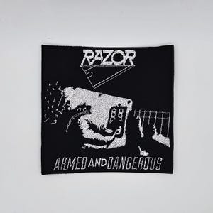 Op de afbeelding: Zwart vierkantige patch met wit geborduurde tekst en afbeelding. Het woord "RAZOR" staat bovenaan, boven een afbeelding van een gitaar. Daaronder staan de woorden "ARMED AND DANGEROUS" geborduurd.