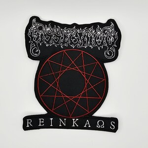 May include: A black embroidered patch featuring a white stylized logo at the top, a red nine-pointed star inside a circle in the center, and the white text REINKAΩS at the bottom. The patch has a black border and a distinct shape with curved edges.