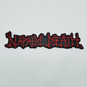 Puede incluir: Un parche bordado rectangular que presenta el texto NAPALM DEATH en una fuente roja estilizada y dentada sobre un fondo negro sólido. Las letras tienen una apariencia espinosa y desgastada, y el parche tiene un borde recortado.