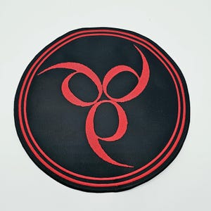 Puede incluir: Un parche circular negro con un símbolo rojo estilizado inspirado en un trisquel en el centro. El diseño está rodeado por dos finos círculos concéntricos rojos cerca del borde exterior, creando un contraste llamativo sobre el fondo negro.