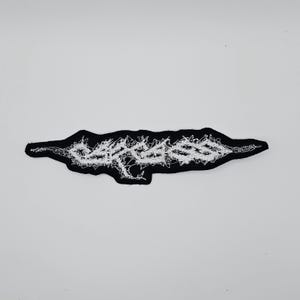 May include: A black embroidered patch with white stitching. The patch has an irregular, elongated shape with a jagged white design in the centre. The patch is set against a white background.