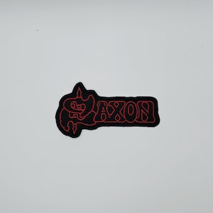 May include: Black and red embroidered patch with the word "AXON" in a stylized font. The patch has a black background with red stitching outlining the letters and a decorative element. The patch is rectangular in shape.