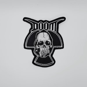 May include: Black and white embroidered patch featuring a skull with a gas mask and the word "DOOM" above it. The patch has a unique shape with a black background and white border.