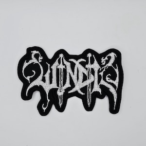 Puede incluir: Un parche bordado negro con la palabra "SUNDER" en blanco, con letras de estilo gótico. Dos espadas se cruzan en el centro de la palabra. El parche tiene un borde negro y está diseñado para coser o planchar sobre tela.