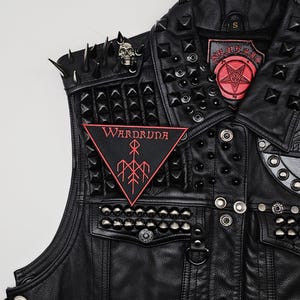 May include: Black leather vest adorned with metal studs, spikes, and patches. Includes a red and black Wardruna patch and a skull pin. The vest has a small size label.