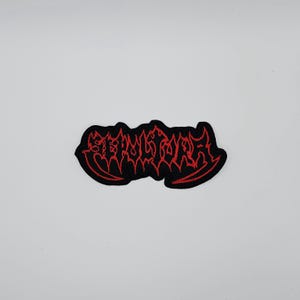 Könnte beinhalten: Schwarzer, gestickter Aufnäher mit dem Wort "STERNUM" in roter, stilisierter Schrift. Der Aufnäher hat einen schwarzen Rand und ist wahrscheinlich zum Aufnähen auf Kleidung oder Accessoires gedacht. Das Design ist auffällig und markant.
