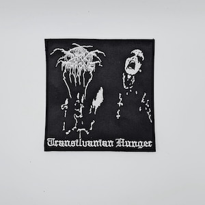Può includere: Toppa quadrata nera con grafica bianca di una figura urlante e forme astratte. Il testo "Transilvanian Hunger" è ricamato in bianco con lettere gotiche. Adatta per abbigliamento o accessori.