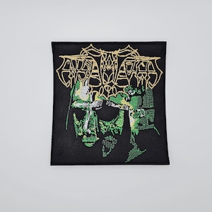 Może przedstawiać: Czarna kwadratowa naszywka ze złotym tekstem i zielono-czarną grafiką. Tekst brzmi "ANTAGONIST". Grafika przedstawia twarz przypominającą czaszkę. Naszywka jest przeznaczona na odzież lub akcesoria.