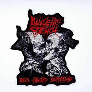 Puede incluir: Parche bordado con un diseño en blanco y negro de dos figuras esqueléticas besándose. El parche tiene texto rojo con la leyenda "INGENIOUS SCENE" en la parte superior y "BEEN CAUGHT ENTERING" en la inferior. Fondo blanco.