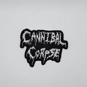 Pode incluir: Patch bordado preto com o texto "CANNIBAL CORPSE" em branco, com letras estilizadas e irregulares. O patch tem uma borda preta e provavelmente é para aplicação em roupas ou acessórios. O design é arrojado.