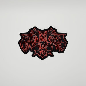 May include: A black embroidered patch featuring a stylized, symmetrical design in bright red thread. The intricate, jagged lines form a complex, abstract shape with sharp points and a dense, web-like pattern throughout the central area.