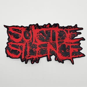 Può includere: Una toppa nera con la scritta "SUICIDE SILENCE" in lettere rosse distorte. La toppa ha un bordo irregolare ed è destinata all'applicazione su abbigliamento o accessori.