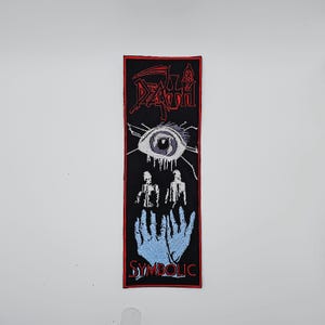 Op de afbeelding: Zwart rechthoekig embleem met rood, wit en blauw geborduurd ontwerp. De bovenkant toont het woord "DEATH" in rood, gevolgd door een oog, twee figuren en een paar blauwe handen. Het woord "SYMBOLIC" staat onderaan.