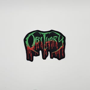 Puede incluir: Parche negro con la palabra "Obituary" en verde, con detalles rojos que simulan goteo de sangre. El parche tiene una forma irregular y es probablemente un parche para planchar o coser en ropa o accesorios.
