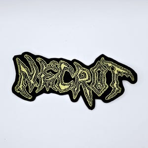 Puede incluir: Parche bordado negro con la palabra "NECROT" en una fuente gótica estilizada. El texto es de color amarillo claro, con un borde negro. El parche tiene una forma irregular.