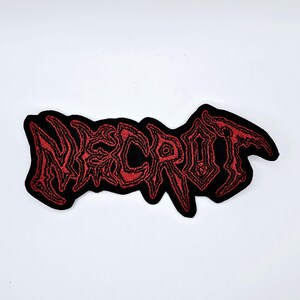 Pode incluir: Um patch bordado preto com a palavra "NECROT" em vermelho, com letras góticas estilizadas. O patch tem uma borda preta e é projetado para ser costurado ou passado a ferro em roupas ou acessórios.
