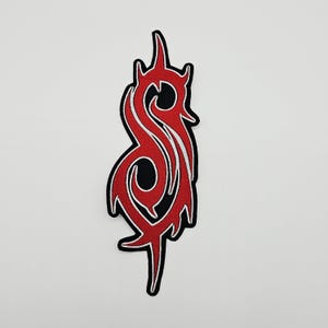 Könnte beinhalten: Ein gestickter Aufnäher mit einem stilisierten, rot-weißen S-förmigen Emblem mit scharfen, spitzen Kanten. Das Design ist mit schwarzem Garn umrandet, was einen starken Kontrast zu den leuchtend roten und weißen Innenbereichen erzeugt.