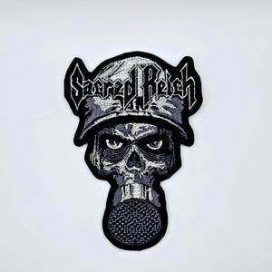 May include: Embroidered patch with a skull wearing a military helmet and gas mask. The patch is predominantly black, white, and grey, with the words "Sacred Reich" in a stylized font across the top.