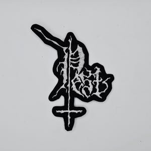 以下が含まれることがあります： 白と黒の刺繍パッチ。骸骨の姿と十字架が描かれています。パッチにはゴシック体のフォントで「RAZOR」の文字がデザインされています。スタイリッシュで詳細なデザインです。