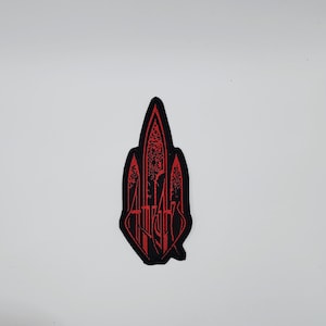Puede incluir: Parche bordado en negro y rojo con un diseño de catedral gótica y texto estilizado. El parche presenta un diseño simétrico detallado con forma de arco apuntado. El texto está en fuente gótica.