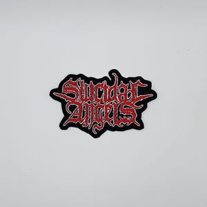 Könnte beinhalten: Ein schwarzer Aufnäher mit dem Schriftzug "Suicidal Angels" in roter, stilisierter, gotischer Schrift. Der Text hat ein zerfleddertes Aussehen und ist schwarz umrandet. Der Aufnäher befindet sich auf weißem Hintergrund.