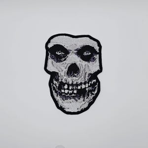 Puede incluir: Parche bordado con un cráneo estilizado con contorno negro. El cráneo tiene un diseño texturizado blanco y gris con cuencas de los ojos, nariz y boca negras. Para ropa o accesorios.