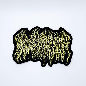 May include: Black embroidered patch with yellow text in a stylized, jagged font. The patch has a black border and is likely designed to be sewn or ironed onto clothing or accessories. The overall design has a heavy metal aesthetic.