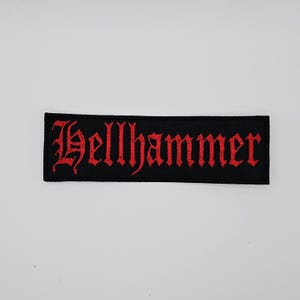 May include: A black rectangular patch featuring the word "Hellhammer" in red gothic script. This embroidered patch is designed for application on clothing or accessories, with a style associated with heavy metal music.