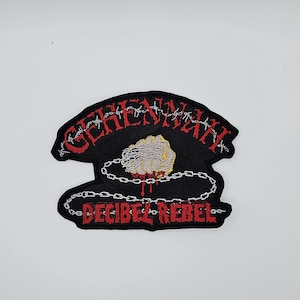 May include: Black embroidered patch with the words "GEHENNAH" in red, barbed wire design, and "DECIBEL REBEL" in red. Features a gray rock with yellow accents, and a silver chain design.
