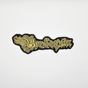 以下が含まれることがあります： 黒色の刺繍パッチ。黄色で「Brosequin」の文字がゴシック様式で描かれています。パッチは黒い縁取りがあり、白い背景に配置されています。衣類やアクセサリーに適しています。