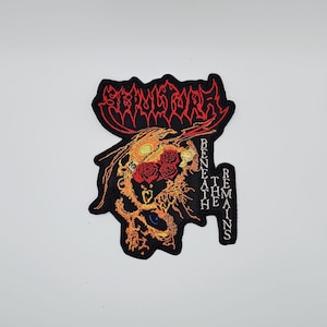 May include: Embroidered patch featuring a skull design with roses and the text "STRANGULATE" in red, and "BENEATH REMAINS" in white. The skull is orange and black, with a blue eye. The patch is black.