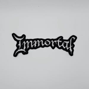 Op de afbeelding: Zwart en wit geborduurde patch met het woord "Immortal" in gotische letters. De patch heeft een zwarte rand en is ontworpen om op kleding of accessoires te worden genaaid of gestreken.