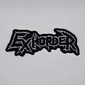 May include: A black patch featuring the word "EXHORDER" in white, jagged lettering. The patch has a black border and is designed to be sewn onto clothing or bags. The style is similar to heavy metal band logos.