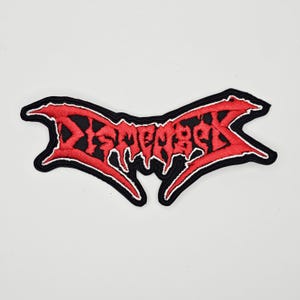 Op de afbeelding: Geborduurde patch met het woord "Dismember" in een rode, gestileerde letter, zwarte rand en witte omlijning. De patch heeft een kenmerkende, gekartelde vorm, ideaal voor kleding of accessoires.