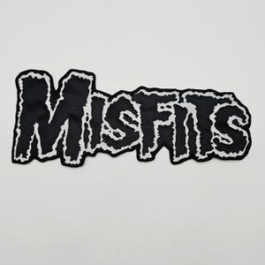 May include: A black fabric patch featuring the word MISFITS in a jagged, stylised font. The lettering is outlined in a thick, textured white border that follows the irregular, distressed edges of each character, creating a high-contrast design.