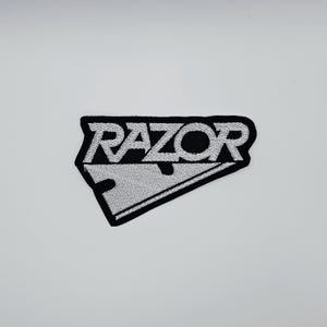 May include: Embroidered patch with the word "RAZOR" in white, stylized text. The patch is black with a white razor blade design. The patch is rectangular with a black border.