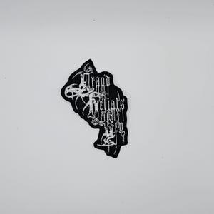 Puede incluir: Parche negro con texto bordado en blanco. El texto es de estilo gótico. El parche tiene una forma única, con una parte superior curva y una inferior puntiaguda. El parche es probablemente para ropa o accesorios.