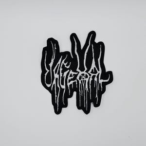 Puede incluir: Parche bordado en blanco y negro con la palabra "Savage Death" en una fuente estilizada y goteante. El parche tiene un fondo negro con costuras blancas para el texto y el diseño.