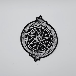 Peut inclure: Écusson brodé noir et blanc représentant un motif circulaire avec une étoile au centre et du texte autour. Le bord est orné d'un serpent qui se mord la queue.