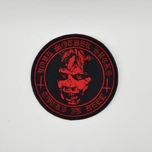 Peut inclure: Écusson rond noir avec texte brodé en rouge et un visage démoniaque. Le texte dit "YOUR MOTHER SUCKS COCKS IN HELL" en police gothique. Deux croix rouges de chaque côté.