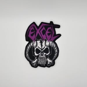 May include: Embroidered patch featuring a skull with horns and crossbones, set against a black background. The word "EXCEL" is embroidered in purple above the skull. The patch is likely for clothing or accessories.