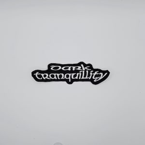Könnte beinhalten: Ein schwarz-weißer Aufnäher mit den Worten "dark tranquility" in einer stilisierten Schriftart. Der Aufnäher hat einen schwarzen Rand und der Text ist weiß. Der Aufnäher befindet sich auf weißem Hintergrund.
