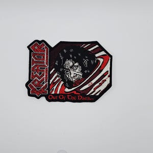 Może przedstawiać: Haftowana naszywka z napisem "KREATOR" w czerwonych, stylizowanych literach. Naszywka ma czarno-czerwony wzór z grafiką potwora i napisem "Out Of The Dark".