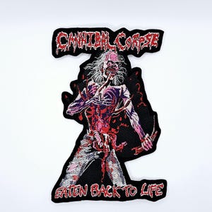 Puede incluir: Parche bordado con un diseño de terror. Representa una figura de tipo zombi con carne y entrañas expuestas. El parche presenta el texto "CANNIBAL CORPSE" encima de la figura y "EATEN BACK TO LIFE" debajo, todo en hilo rojo.