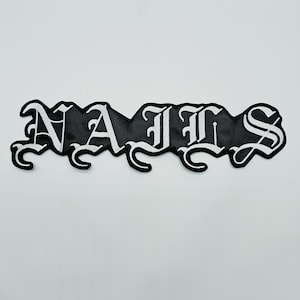May include: A black embroidered patch featuring the word NAILS in a white gothic-style font. The letters are connected in a single horizontal line with decorative flourishes at the base of each character, creating a bold, high-contrast design.