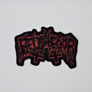 Puede incluir: Parche negro con texto rojo y símbolos de cruz. El texto parece ser el nombre o logotipo de una banda. El parche tiene un borde negro y probablemente está hecho de tela.