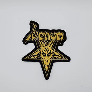 May include: Black and gold embroidered patch featuring the word "Venom" in a stylized font above a gold pentagram with a goat's head. The patch is likely for a jacket or bag.